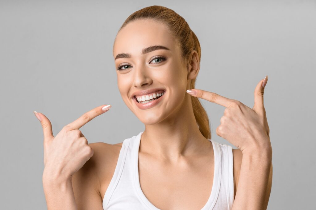 A smiling woman in a white tank top pointing at her bright, white teeth against a plain gray background.